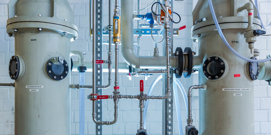 Two light gray containers connected with different pipes made of metal stand next to each other in the laboratory.
